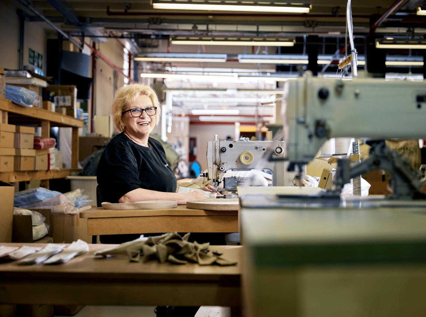 A blonde woman wearing glasses smiling at the camera as she works at a sewing machine