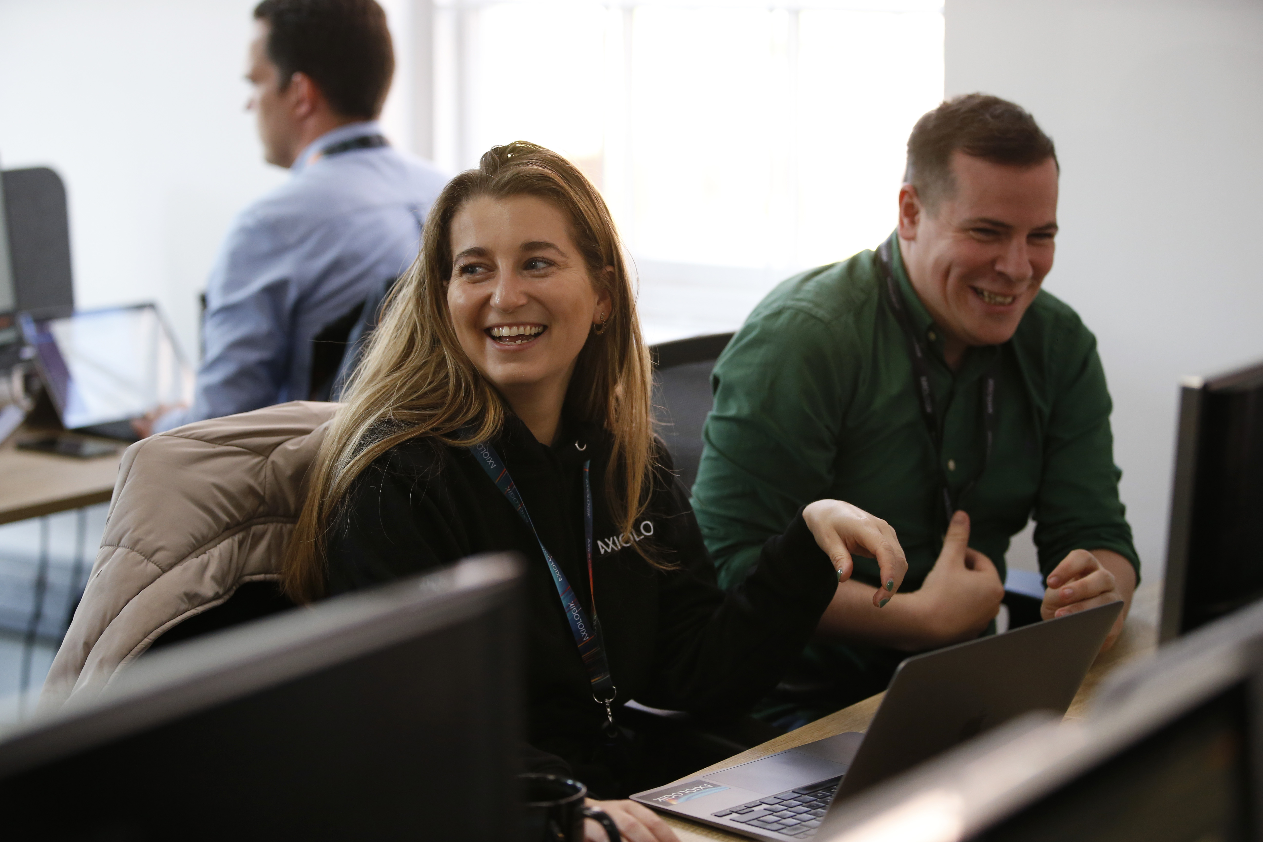 Two members of the Axiologic team are showing laughing in their office whilst sat at their desks