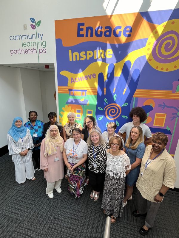 A group of women are shown smiling for a camera positioned above them, Engage, Inspire, Achieve is shown on a colourful mural behind them