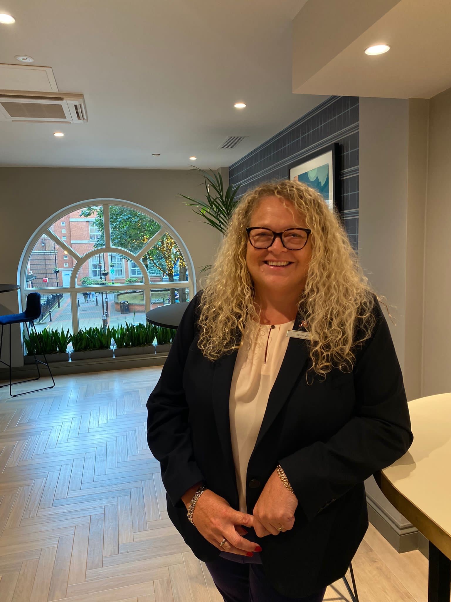 Jayne from Leeds Marriott Hotel smiles for the camera, she is leaning against a desk within the hotel