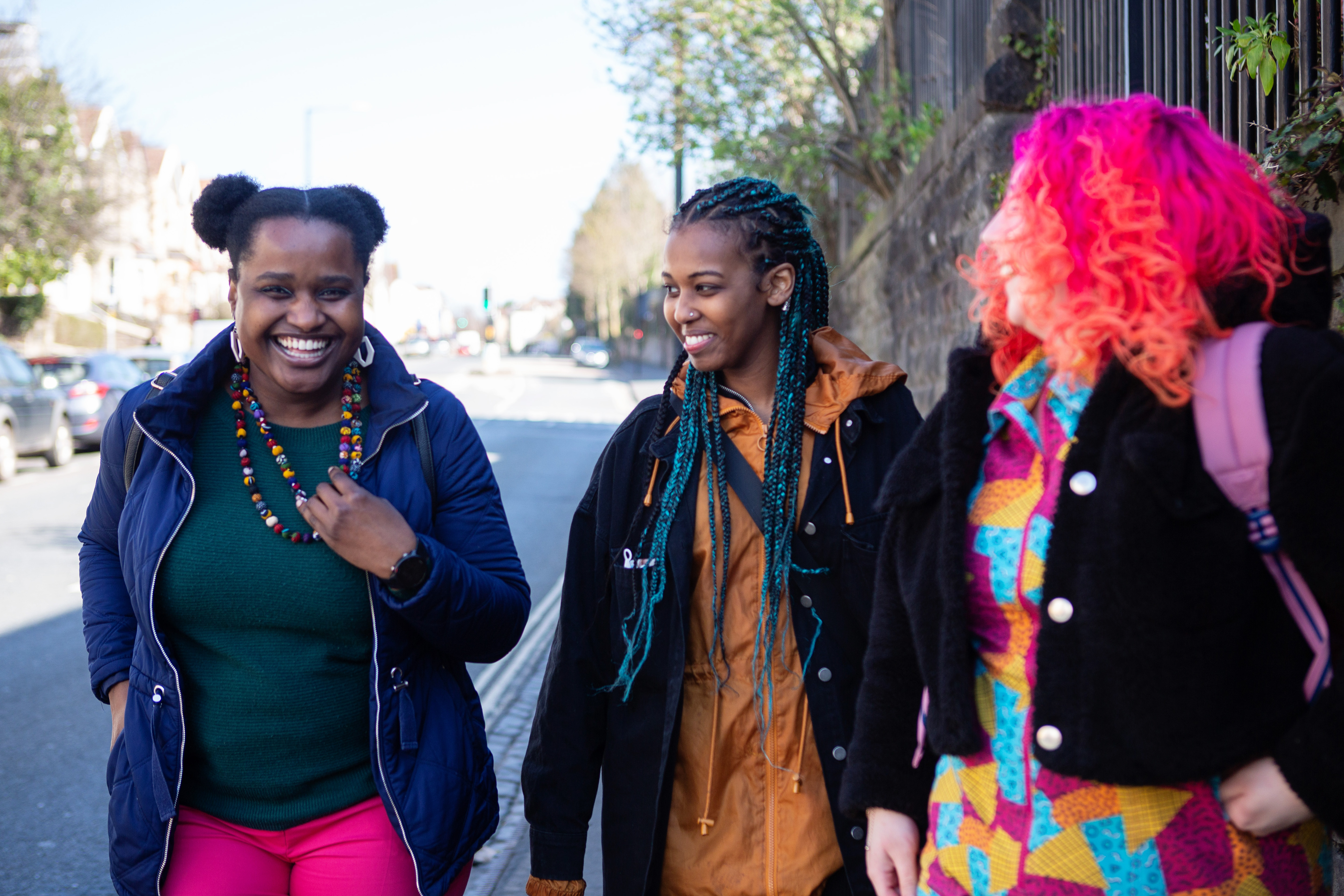 Two black women and one white woman are walking through Leeds, all three are wearing colourful clothes and laughing as they walk