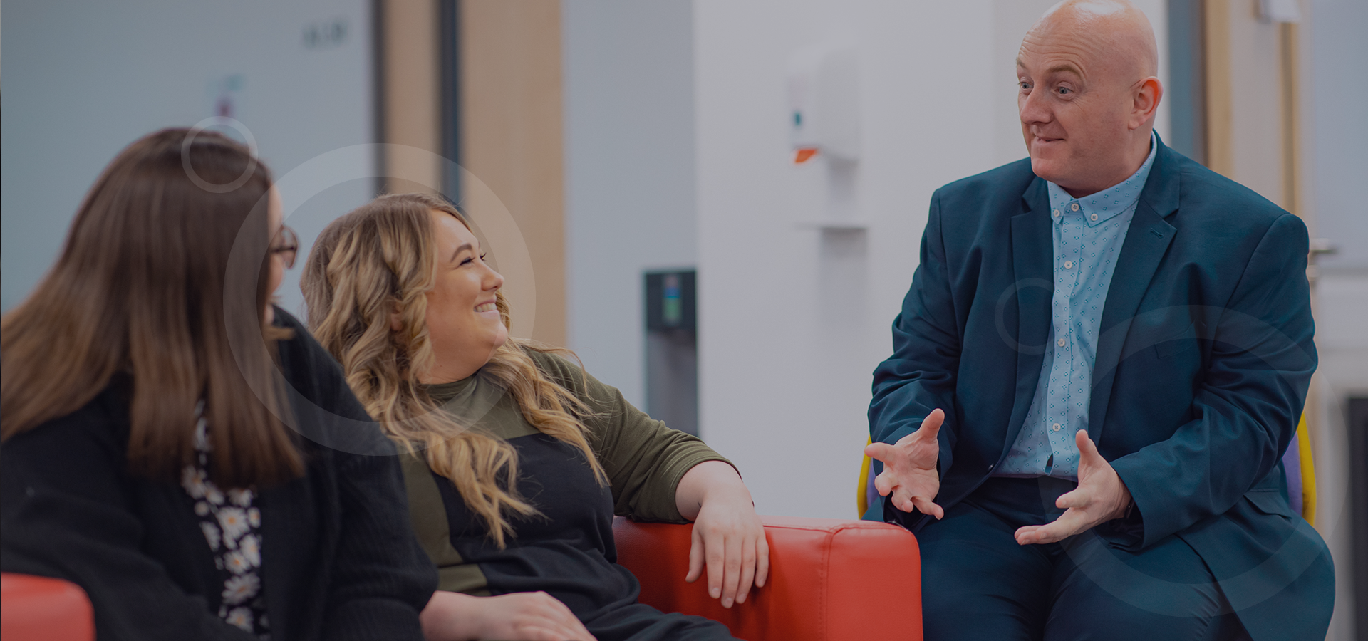 Two women sat on a sofa talking with a man sat on a chair.
