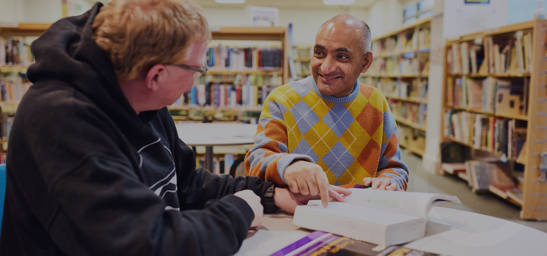 A man pointing to a page in a book with another man whilst they sit in a library.