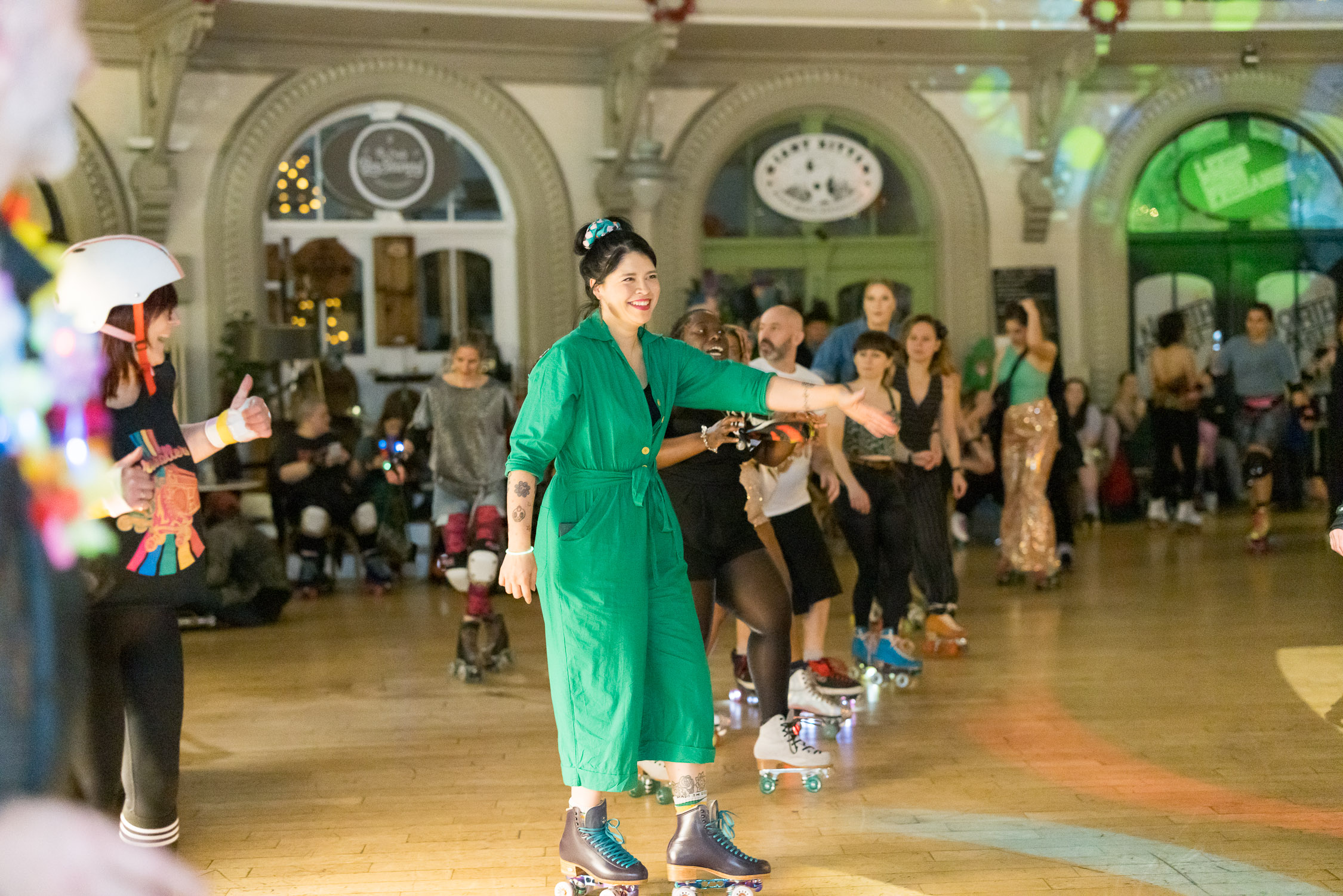 A lady in a green jump suit is shown wearing roller skates, behind her is a group of other skaters