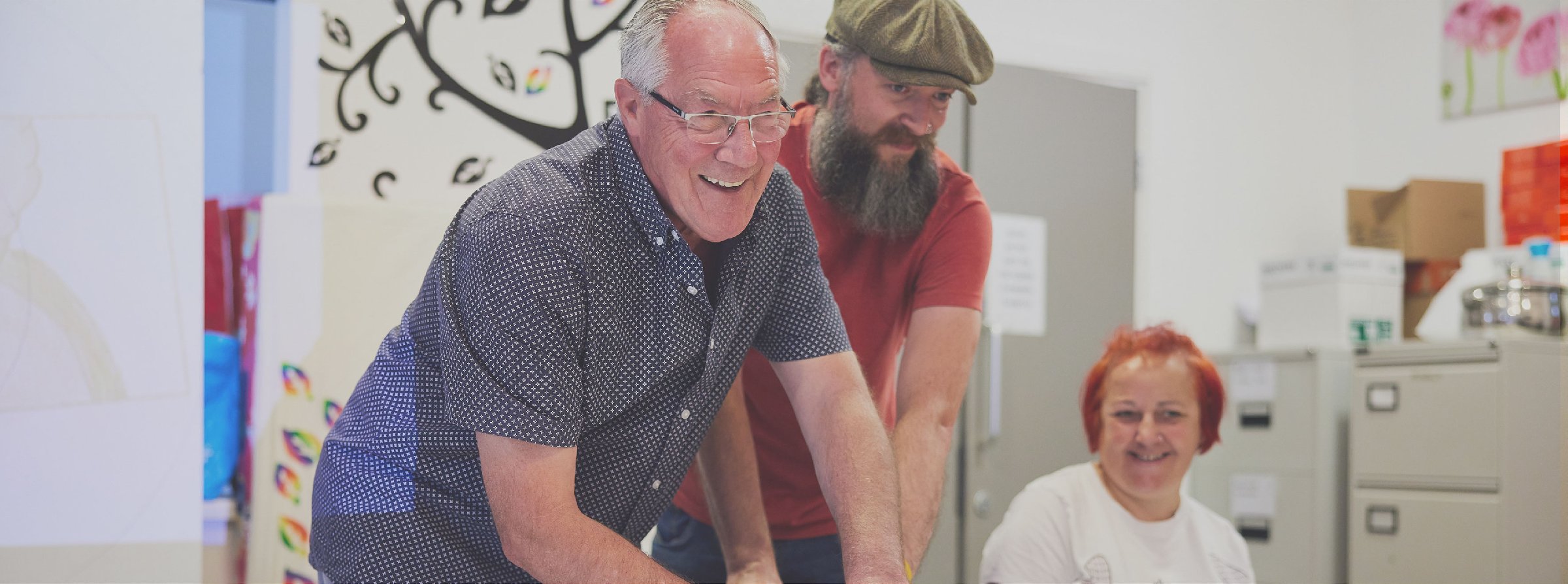 A man wearing glasses, another man wearing a flat-cap and an orange haired woman sitting down smiling together.