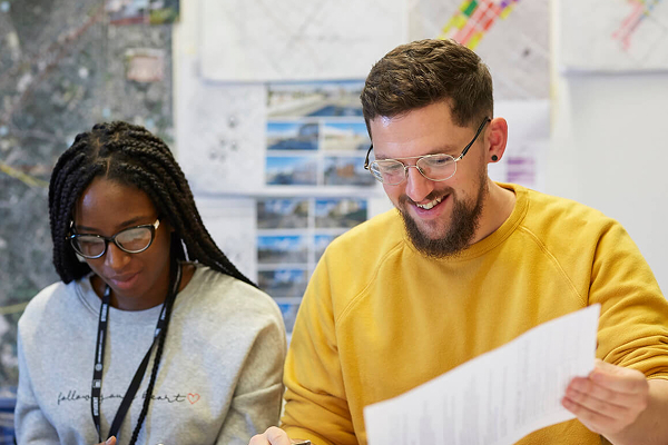 A black female student and white male student are shown sat together smiling whilst looking at paperwork