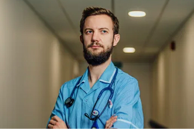 A male doctor in blue scrubs is looking down a corridor with his hands folded